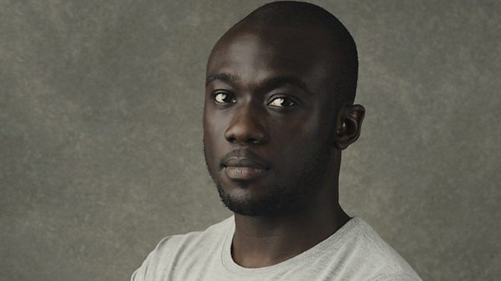 Segun Akinola wears a grey shirt in front of a grey wall.