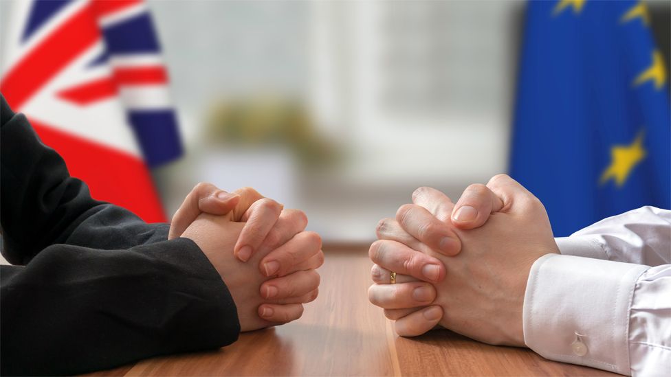 Generic image of two men's hands clasped at a negotiating table, with the Union Jack and EU flag behind them