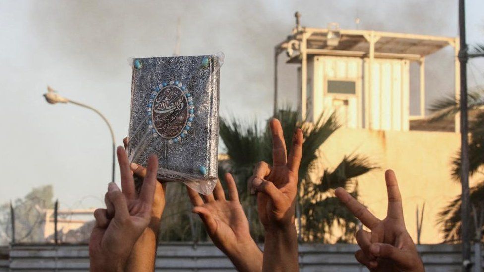 A protester holds up a Koran as smoke rises from the Swedish embassy building as protesters gather near the embassy in Baghdad hours after it was stormed and set on fire ahead of an expected Koran burning in Stockholm