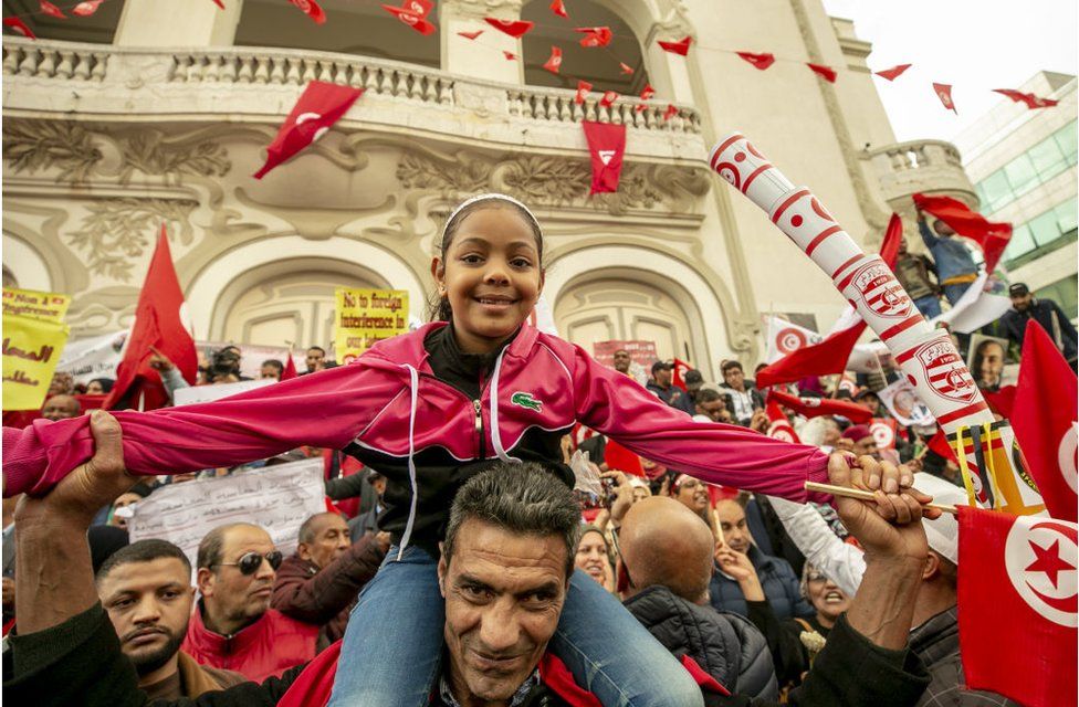 Girl with her arms stretched out wide smiling and celebrating amongst a cheerful crowd who have Tunisian flags in their hands.