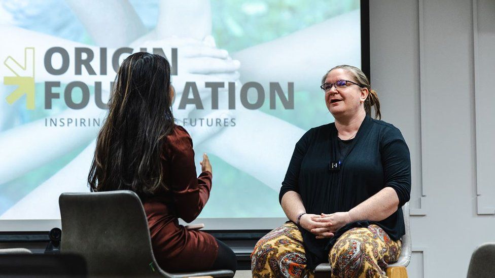 Two women sat on stage talking with each other, in the background there is a presentation slide3 that says 'Origin Foundation'