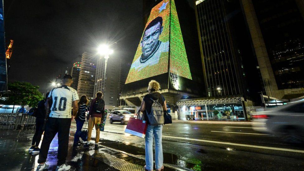 People stand on a pavement across the road from a building lit up with the face of Pele