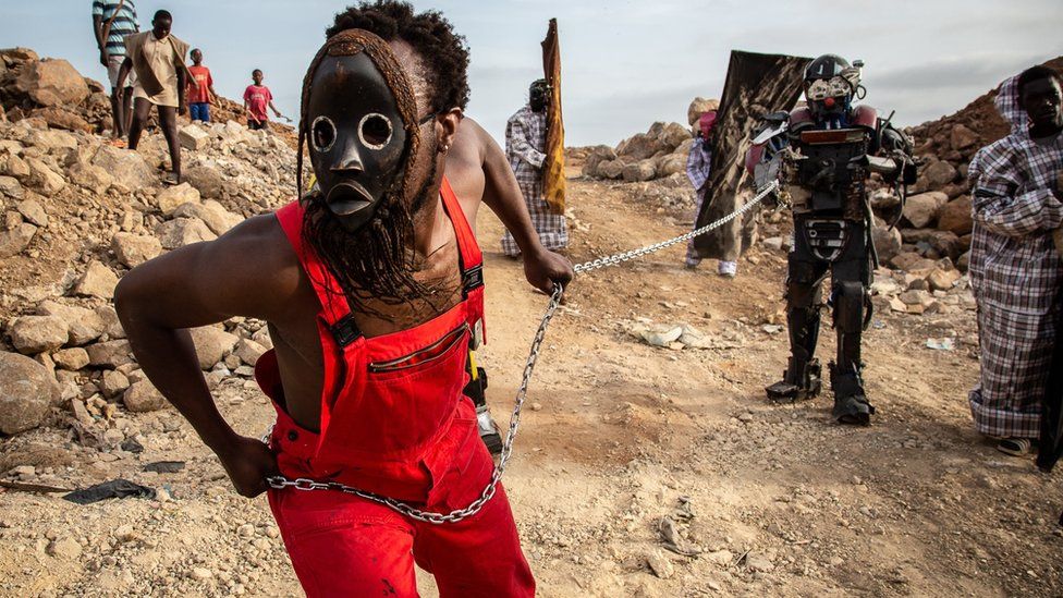 A performance takes place along the coastal walkway on May 21, 2022 as part of the Dakar Biennale. The performer is wearing a mask and is holding a chain, which is attached to another performer.