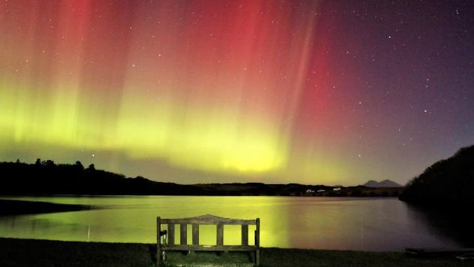 Bench over a body of water with red and yellows of the aurora reflected.
