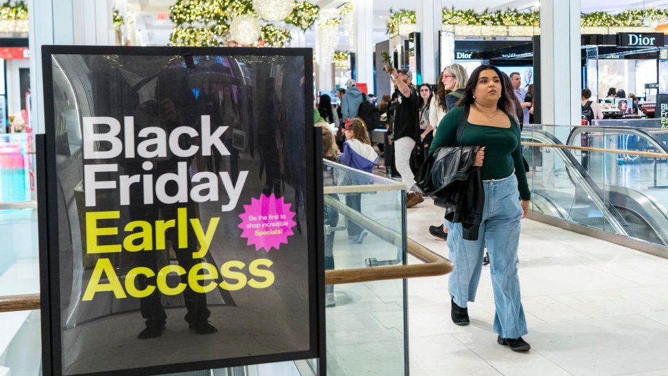 Black Friday advertising in a shopping centre - generic image. A black poster reads 'Black Friday Early Access' in white and yellow font. A woman with long dark hair wearing a green top and denim jeans walks past. In the background are other shoppers and outlets and an escalator