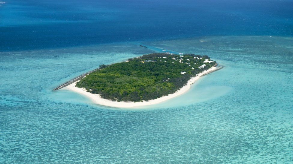 A small island with sandy beaches and trees, surrounded by a coral reef