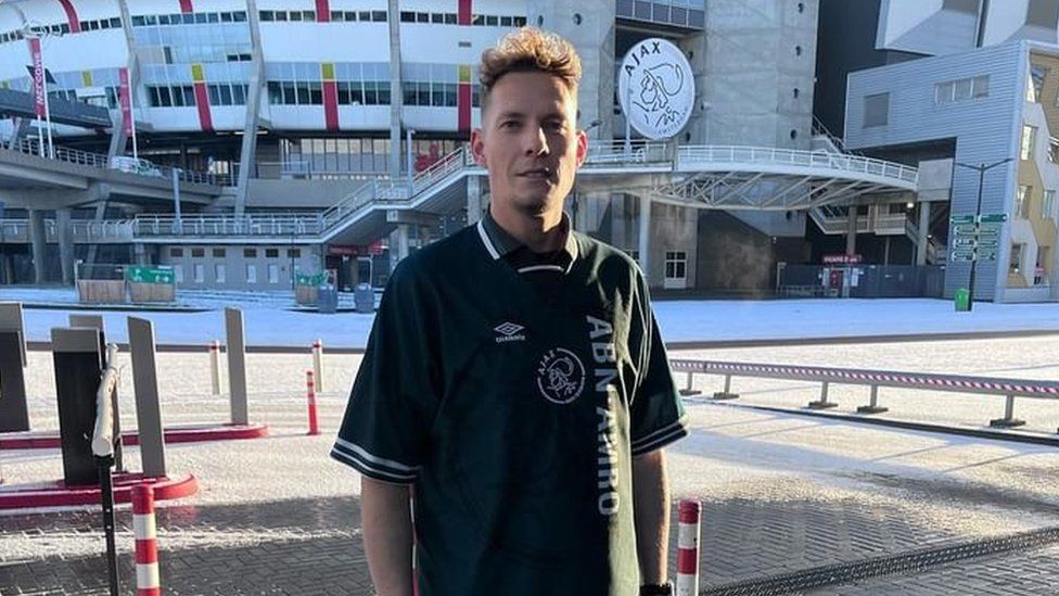 Scott Stuurman pictured outside the Ajax stadium in Amsterdam. Scott is a white man with curly brown hair and brown eyes. He wears a green Ajax polo shirt despite there being snow on the ground outside the stadium behind him.
