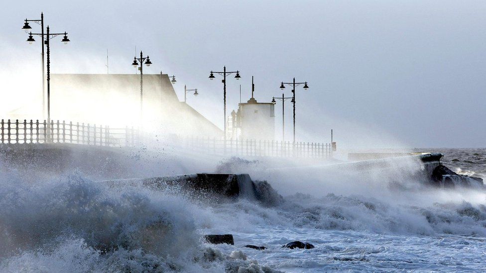 Waves breaking over sea wall with houses behind