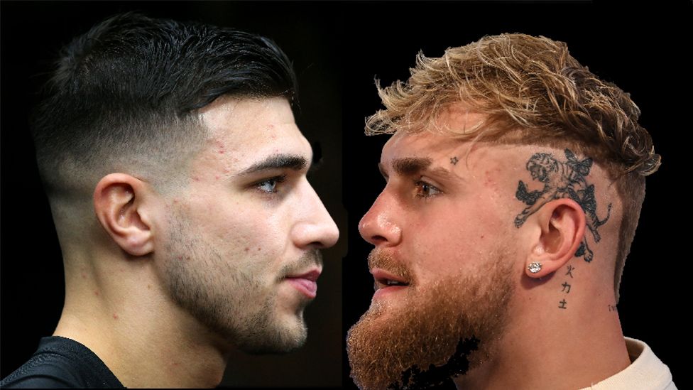 Tommy Fury, a white man with short, dark hair, blue eyes and a short stubble faces off against YouTuber Jake Paul, a white man with wavy, sandy blonde hair, brown eyes and a beard. Paul has a number of tattoos including a star above his eyebrow and a tiger with butterflies above his left ear and also has a diamond earring.