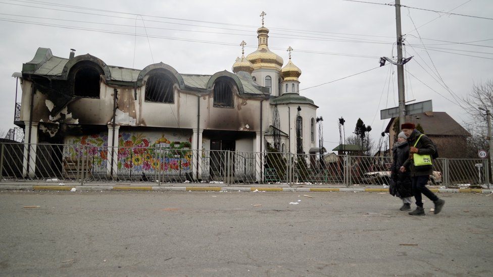 A smoke-darkened, burned out building with a seemingly undamaged Ukrainian church behind it, complete with three golden onion domes