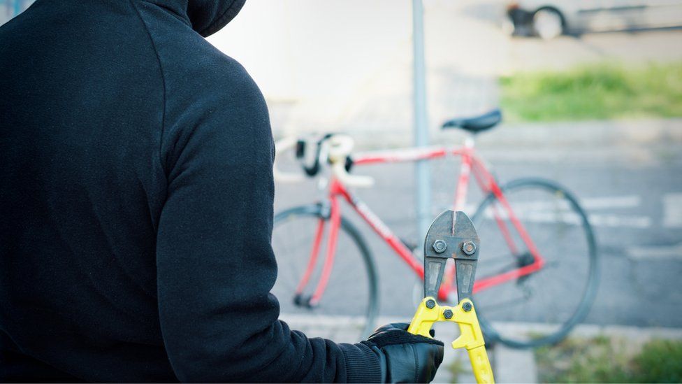 Hooded and gloved thief holding bolt cutters approaches a bike which is locked against a post