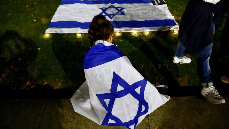 A woman draped in an Israel flag sits before another flag surrounded by candles at a vigil in Downing Street, October 2023