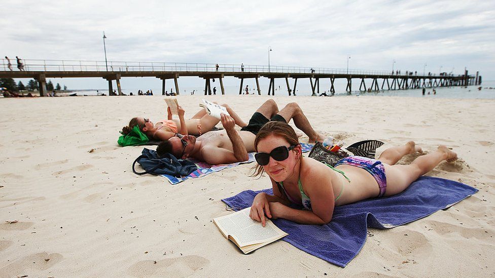 Две женщины и мужчина лежат на пляже в Гленелге в Южной Австралии.
