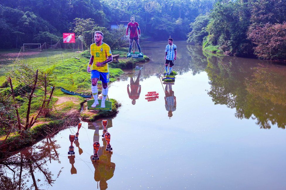 Fans wave flags next to the giant cutouts of players from Brazil's Neymar (L), Portuguese Cristiano Ronaldo (C) and Argentine Lionel Messi, erected by football fans in river Cherupuzha at Kozhikode in India's Kerala state on November 7, 2022, ahead of the Qatar 2022 FIFA World Cup football tournament.