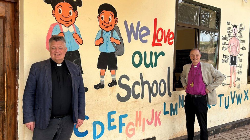 Reverend Mark Bee and Bishop Mike Harrison stand in front a mural on an African school wall with images of students and colourful writing