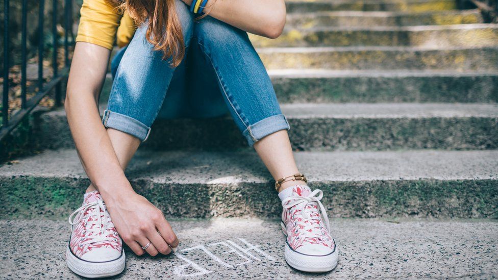 A teenage girl uses chalk to write the word "help" as she sits on a step