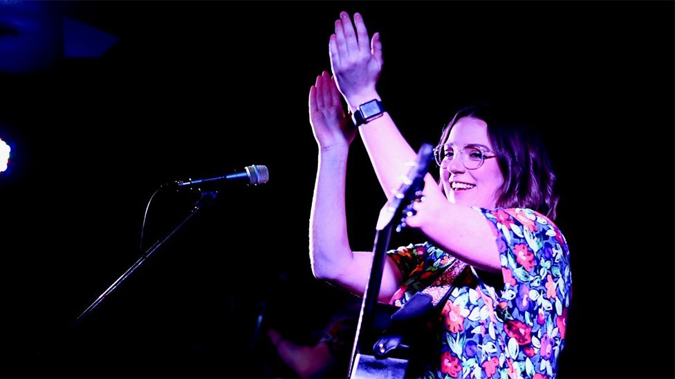 Lucy is wearing a floral top, with a guitar around her, clapping as she sings into the two microphones in front of her. She is wearing a watch on her left wrist. The background is plain black.