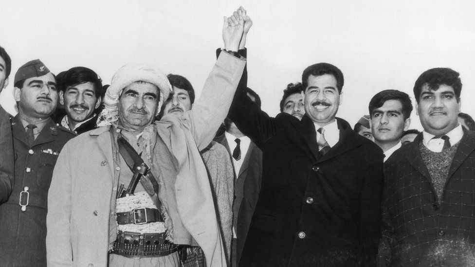 Mulla Mustafa Barzani, líder do Partido Democrático do Curdistão, de mãos dadas com Saddam Hussein, então vice-presidente do Conselho de Comando Revolucionário do Partido Baath iraquiano (20 de março de 1970)