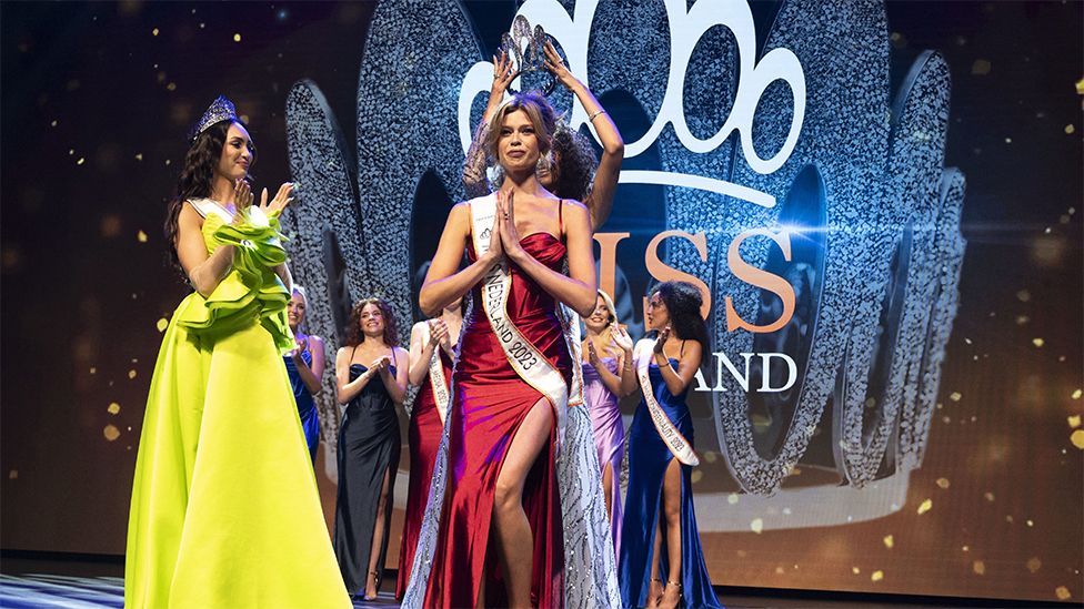 Rikkie Valerie Kolle being crowned Miss Netherlands on stage. She is wearing a red dress and has a white sash around here around here saying Miss Netherlands 2023. She has a big silver tiara on her head and is surrounded by other Miss Netherlands competitors wearing similar dresses and smiling and clapping