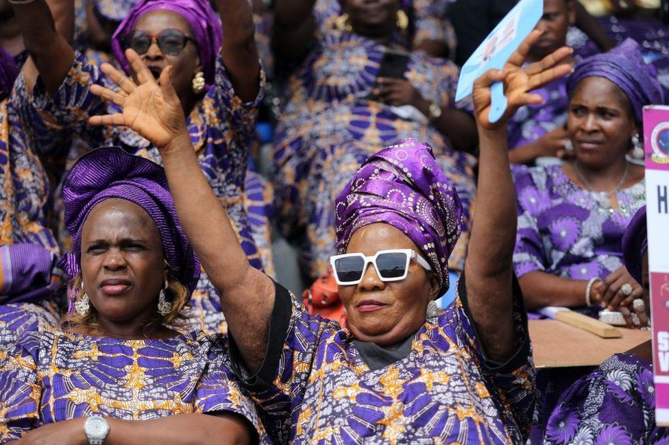Women wearing traditional Nigerian clothes in purple and yellow. There is a woman in the middle wearing white-framed sunglasses who is raising her hands in the air.