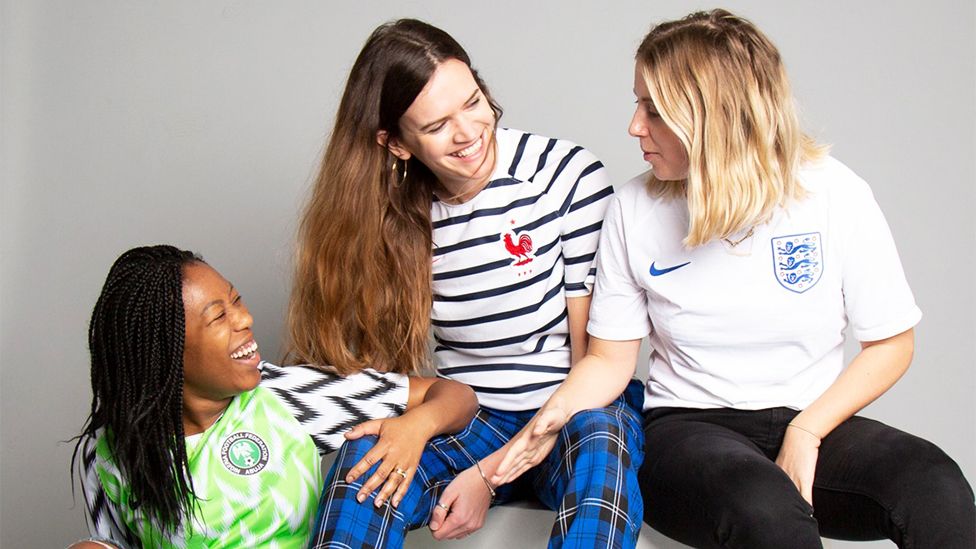 Amy está en el centro, con dos personas, una a cada lado. Amy sonríe y lleva un top a rayas blancas y negras con pantalones de tartán a cuadros azules y negros. La señora de la izquierda está agachada y lleva una camiseta de fútbol verde y blanca, mientras que la señora de la derecha lleva una camiseta blanca de Inglaterra con pantalones negros.