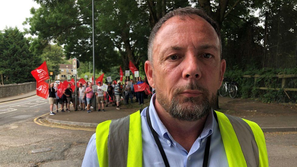 Ken Fish wearing a fluorescent jacket with striking Unite workers in the background