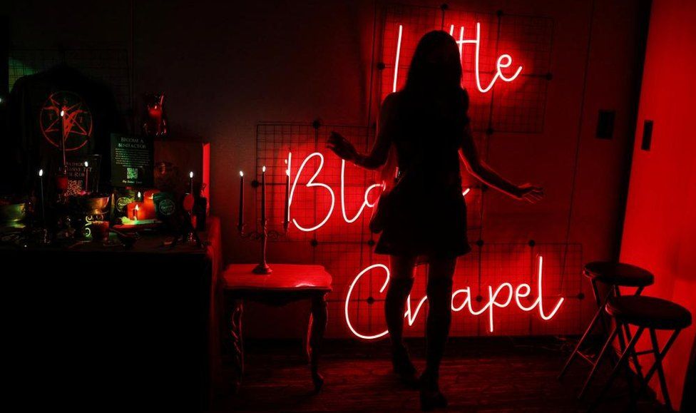 An attendee poses for a photograph inside the Little Black Chapel at The Satanic Temple's Satancon 2023. The figure is silhouetted against a wall lit by a red neon sign reading 'Little Black Chapel'. A table with candles on it stands to the left.