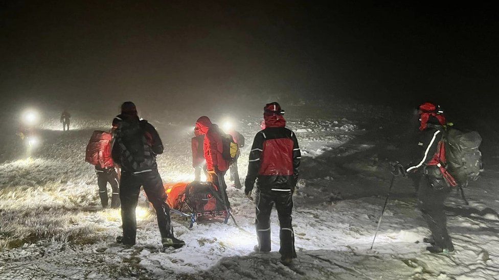 Volunteers in red and black coats and lit by headtorches push a stretcher across snow and rocks