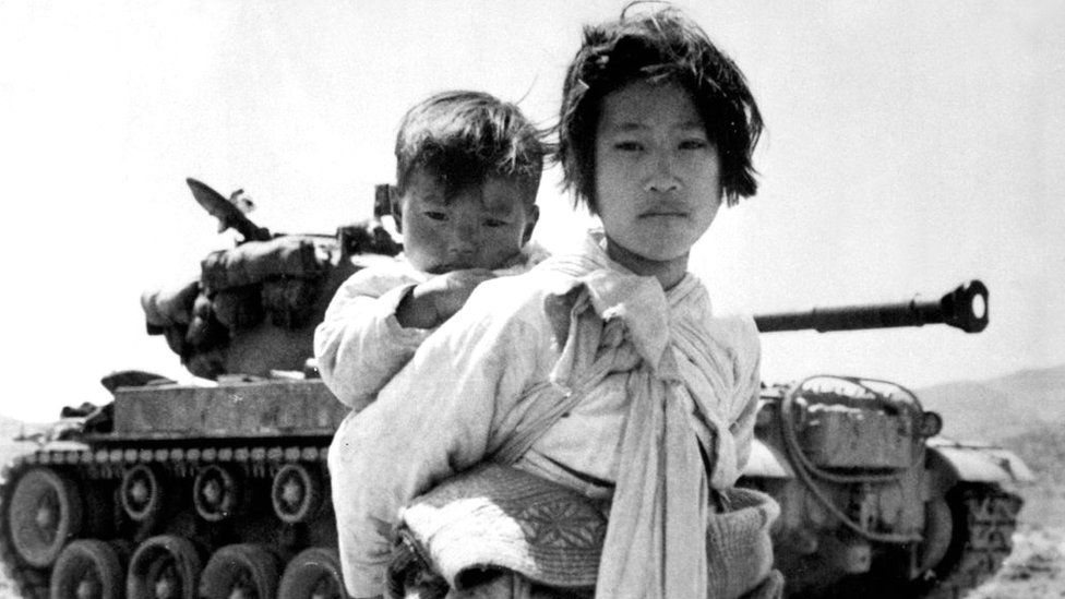 With her brother on her back a war-weary Korean girl trudges by a M-26 tank at Haengju.