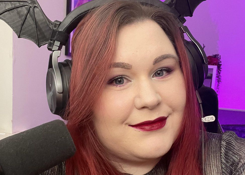Streamer Nic wears a dark grey gaming headset with plastic bat sticking out of the top on either side. She's got reddish brown hair and wears a deep red lipstick.