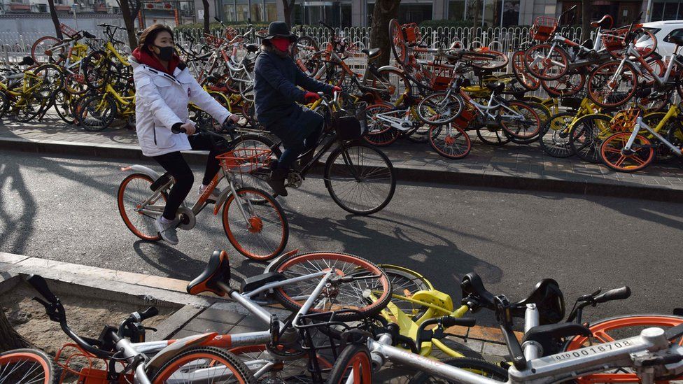 People ride past shared bicycles piled beside a road in Beijing in 2018.