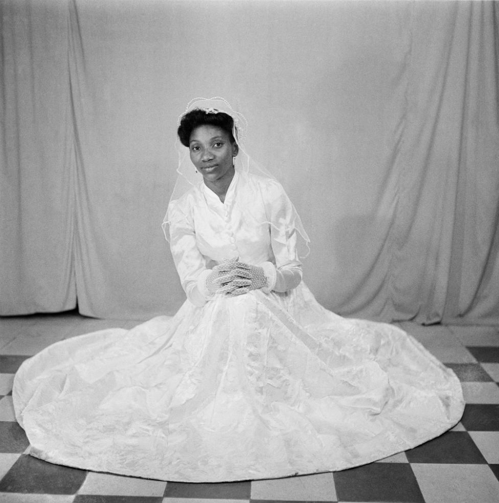 A woman poses on the studio floor in a white wedding gown, lace gloves and veil.