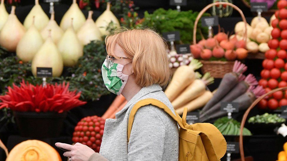 A woman in a face mask walks past an arrangement of vegetables.