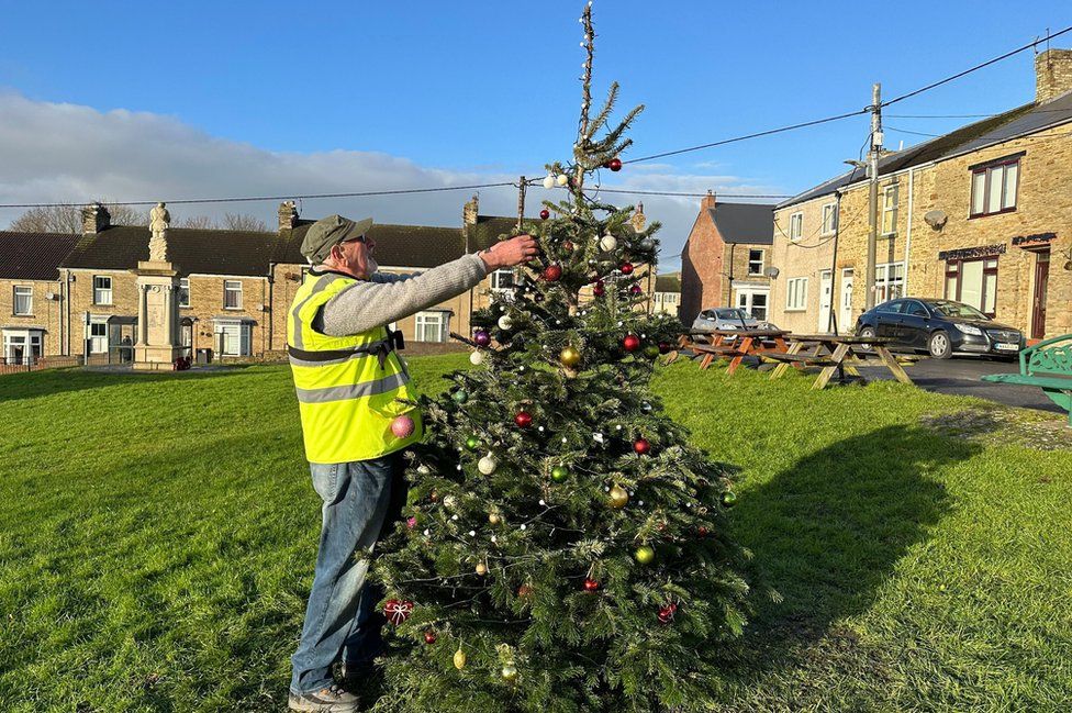 A man moves a bauble on a Christmas tree on a village green