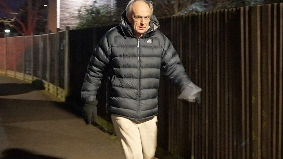 Man wearing gilet walking down a street with a fence on his left
