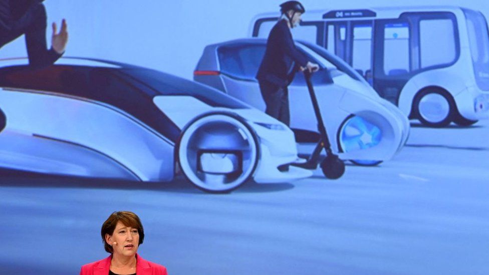 Hildegard Mueller stands before a graphic showing electric powered cars, scooters and busses