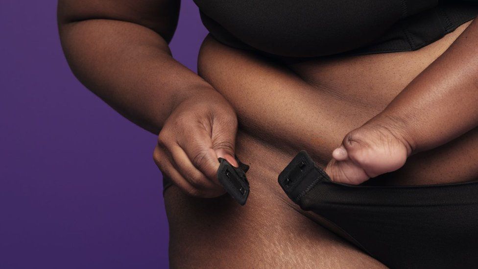 A shot of the accessible underwear worn by a model. A black woman with a hand disability holds the briefs, which have magnetic clasps on the hips, together as if to connect them. The underwear is black and only the model's arms and midriff is photographed against a purple background.