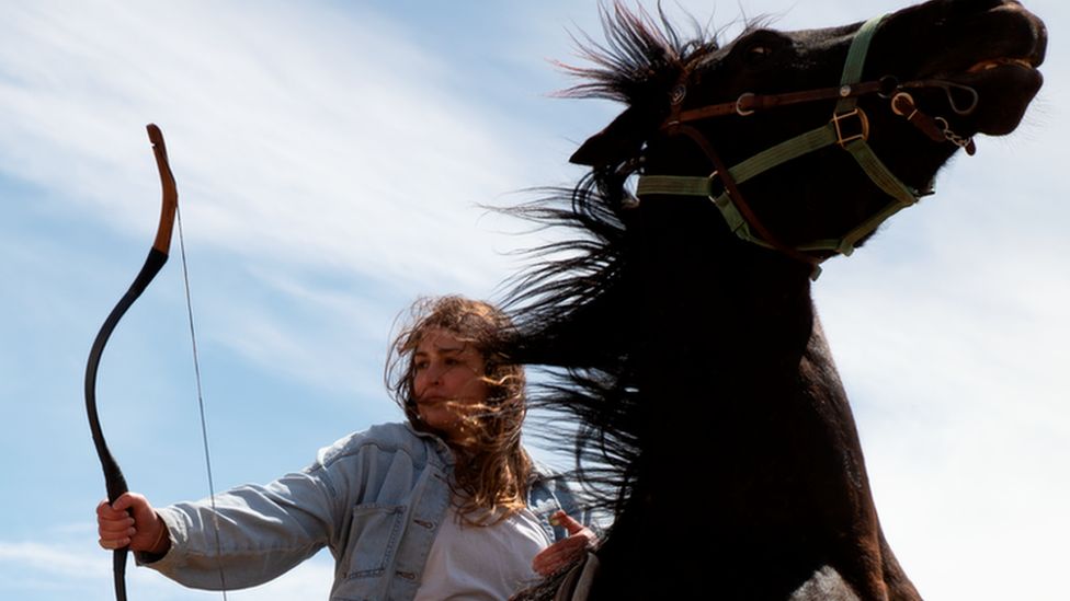 Catriona Scott, a 30-year-old white woman with long brown hair, rides a black horse as it rears up. She holds a bow in her right hand, looking to the side. Catriona wears her hair loose and it's caught in the wind along with the horse's mane. She wears a denim jacket, jeans and a white top. She's pictured outside on a sunny day against a blue sky dusted with clouds.