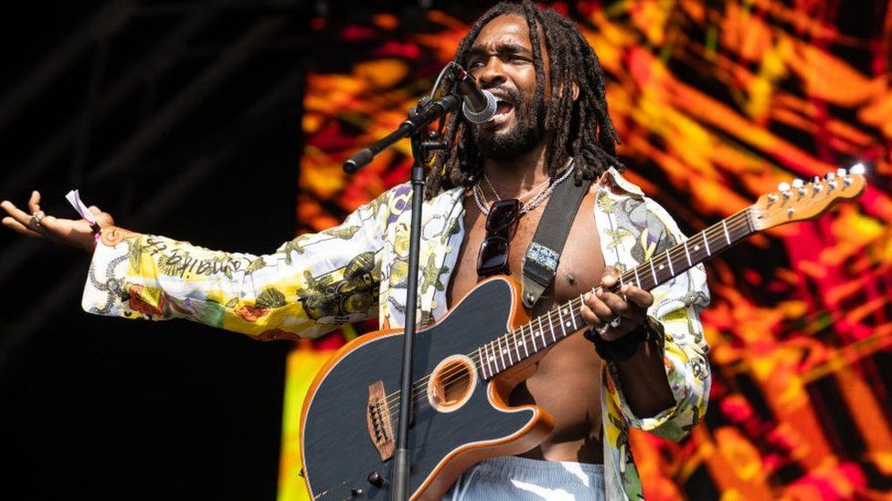 Hak Baker performing on stage. He is singing and holding a black guitar in front of a mic. He has shoulder length dreadlocks and is wearing a patterned shirt that is unbuttoned all the way down. His left hand is on his guitar neck and his right arm is outstretched to his side