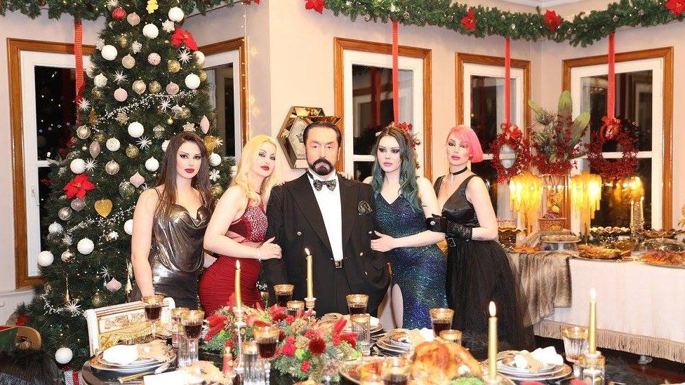 Adnan Oktar stands in a room decorated for Christmas, surrounded by four women on his arms with varying dyed hair colours