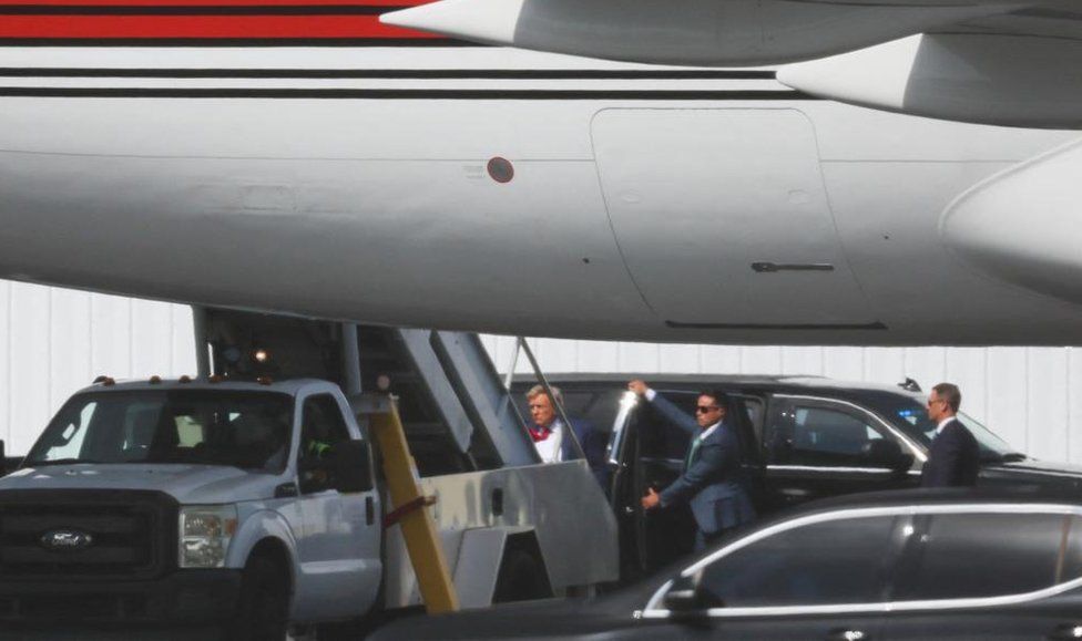 На фото Дональд Трамп поднимается по лестнице своего самолета, чтобы покинуть Майами, а помощник держит открытой дверцу машины на взлетно-посадочной полосе позади него