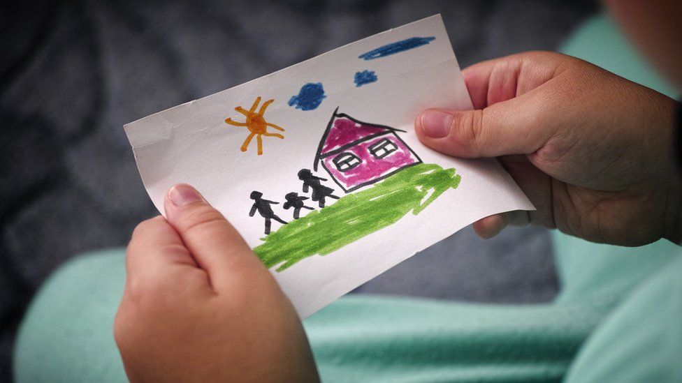 Child holding a children's drawing of a house and stick man, woman and child