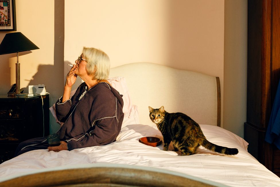 A woman sits on the bed smoking with her cat beside her