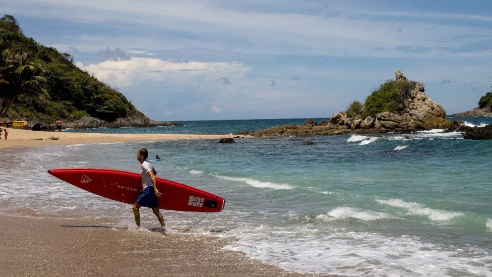 A man carrying his paddle board out of the water on a beach in Phuket