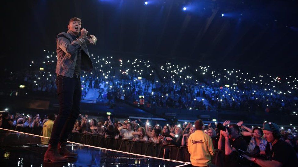 Man with short dark hair sings into a microphone on a stage. Audience holding lights.