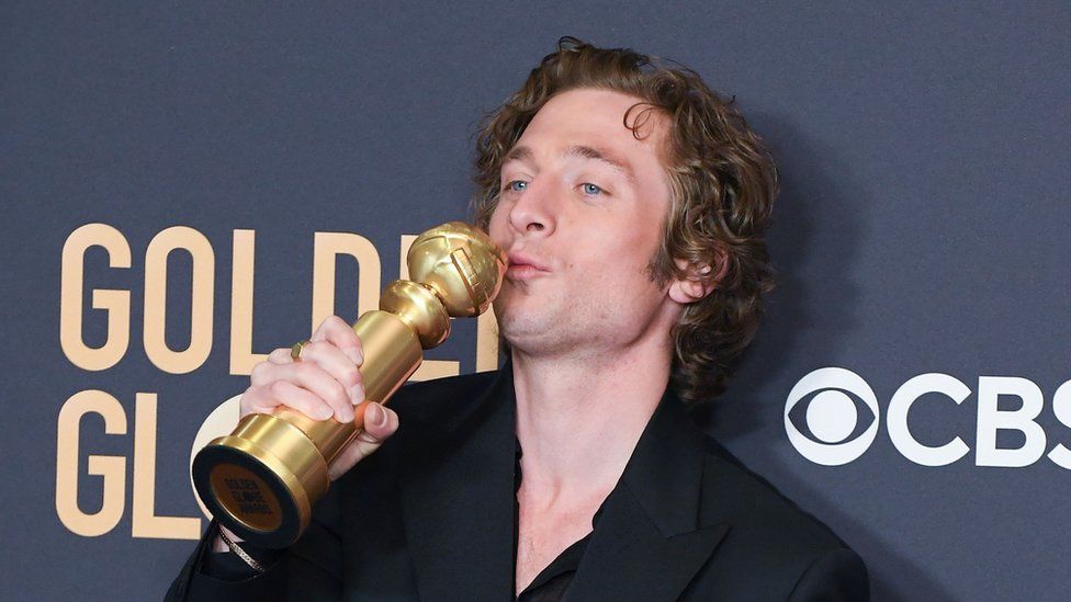 Jeremy Allen White, a white man with short, wavy hair, kisses a Golden Globe award statue as he stands in front of a red carpet hoarding with "Golden Globes" and "CBS" logos printed on it. He's wearing a black suit jacket and black dress shirt, unbuttoned to the chest.