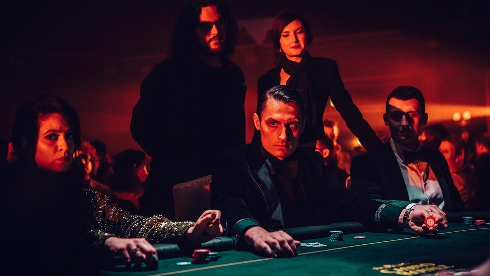Players sit at a card table in costume, with the main villainous actor, blinded in one eye, starting at the camera - an example of the productions that go on around the film