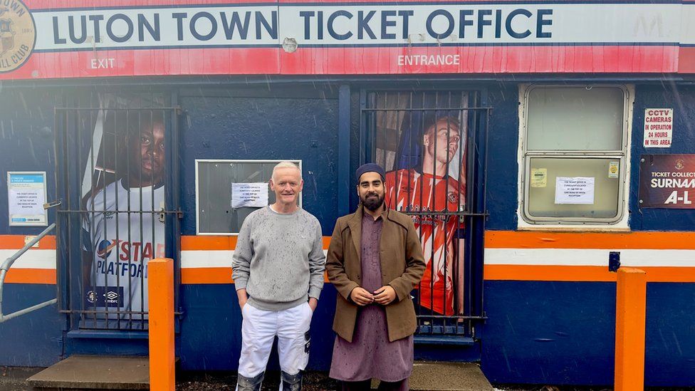 Former EDL member Darren Carroll and Imam Dawood Masood stood with the Luton Town ticket office entrance behind them. Darren is on the left and is stood with his hands in his pockets wearing white trousers and a grey jumper which looks like it has rain on it. He also has short grey hair. Dawood is stood to his right directly next to him and is wearing a purple kameez (a long shirt) and a brown coat over the top with a navy kufi (cap) on his head.