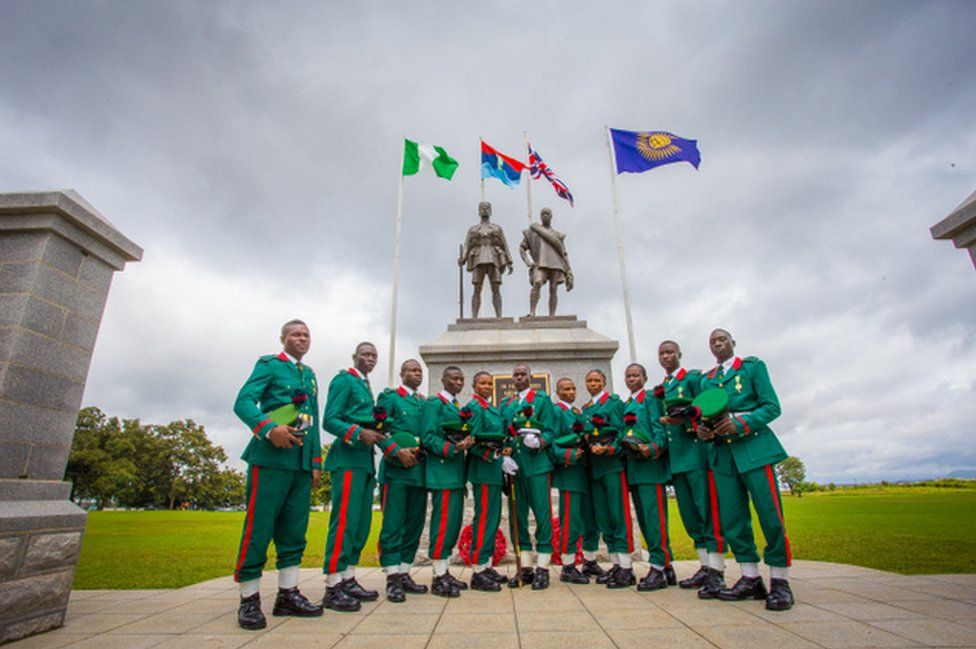 Male and female soldiers in green and red uniforms stand in front of the memorial featuring flags and two statues in Abuja