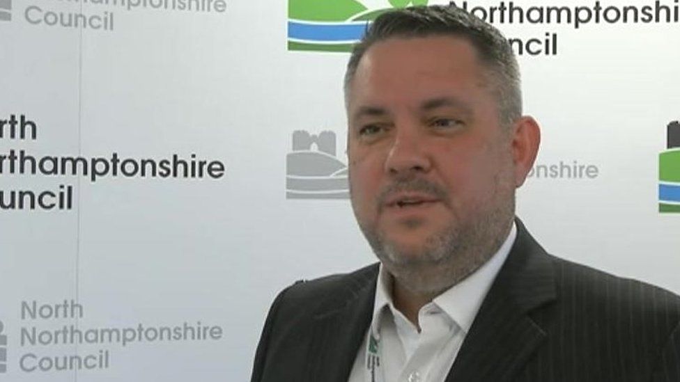 Man with short dark hair and faint beard wearing a jacket in front of North Northamptonshire Council signs